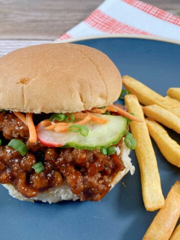 Sandwich sits on a dark blue plate with a red and white striped napkin under it. Top bun is slightly askew to show the sloppy joe topped with pickled vegetables. To the right of the sandwich is a pile of french fries.