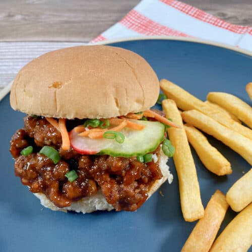 Sandwich sits on a dark blue plate with a red and white striped napkin under it. Top bun is slightly askew to show the sloppy joe topped with pickled vegetables. To the right of the sandwich is a pile of french fries.