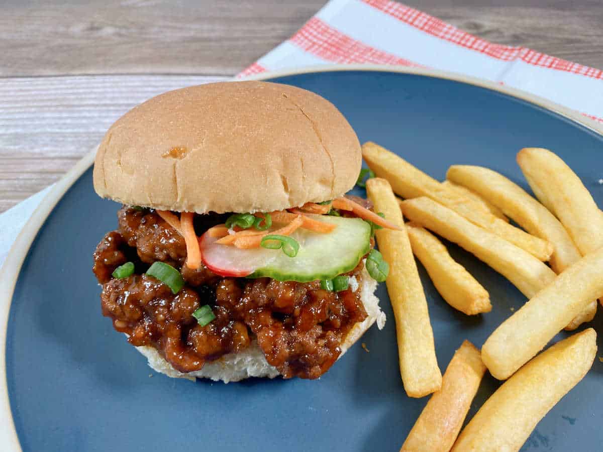 Sandwich sits on a dark blue plate with a red and white striped napkin under it. Top bun is slightly askew to show the sloppy joe topped with pickled vegetables. To the right of the sandwich is a pile of french fries.