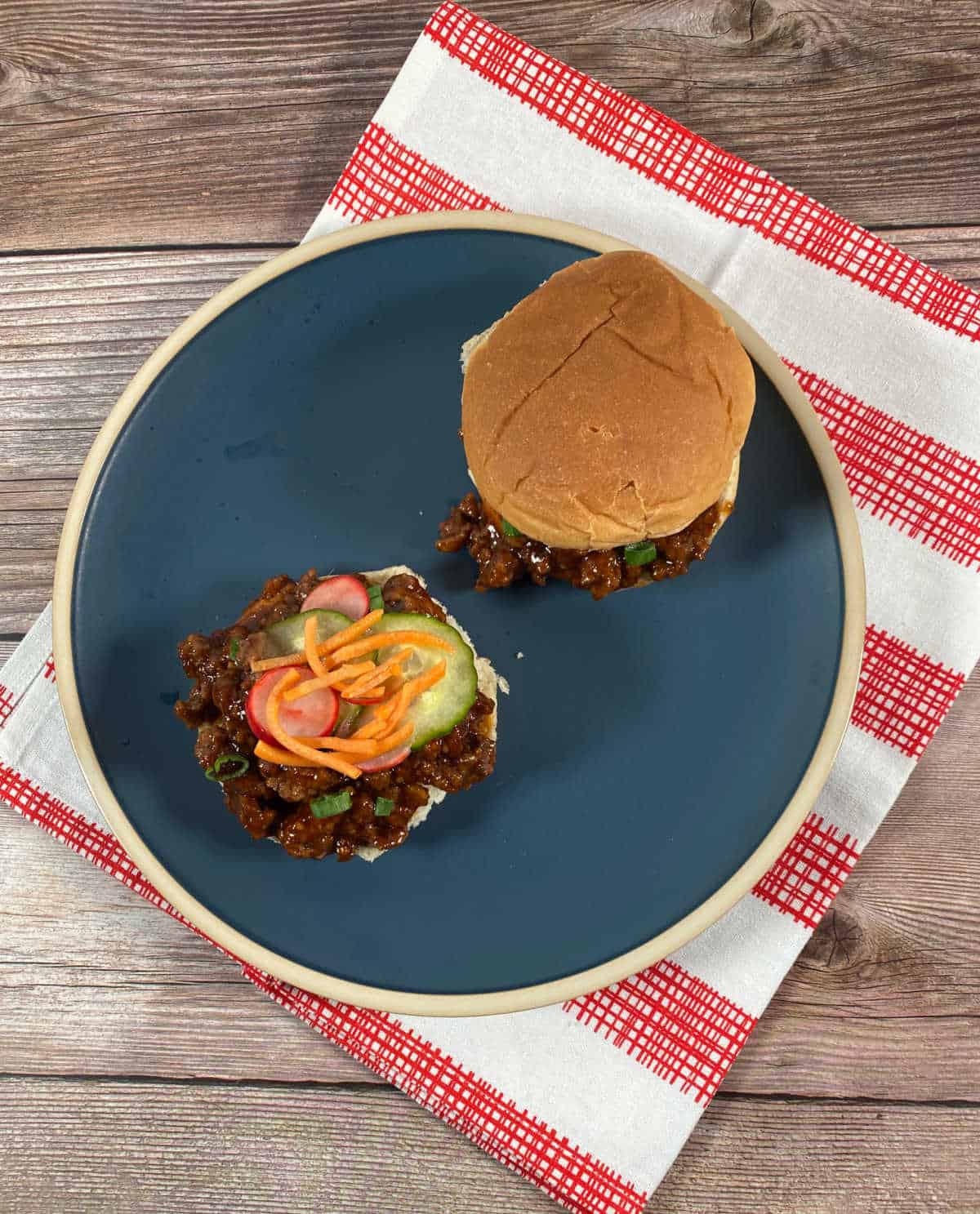 Overhead image of sandwiches on a dark blue plate under a red and white striped napkin. One sandwich is fully assembled and the oter has the top bun off, showing the pickled vegetables resting on the meat and the diced green onion mixed in. 