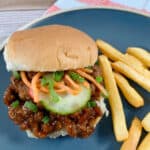 Sandwich has bun slightly askew to show pickled vegetables on top. Sandwich sits on a dark blue plate with fries to the right of it. A red ad white striped napkin sits in the background.