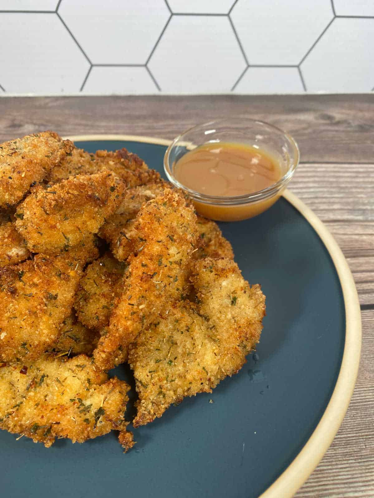 Chicken tenders piled up high on a blue plate with a glass dish of honey mustard for dipping. 