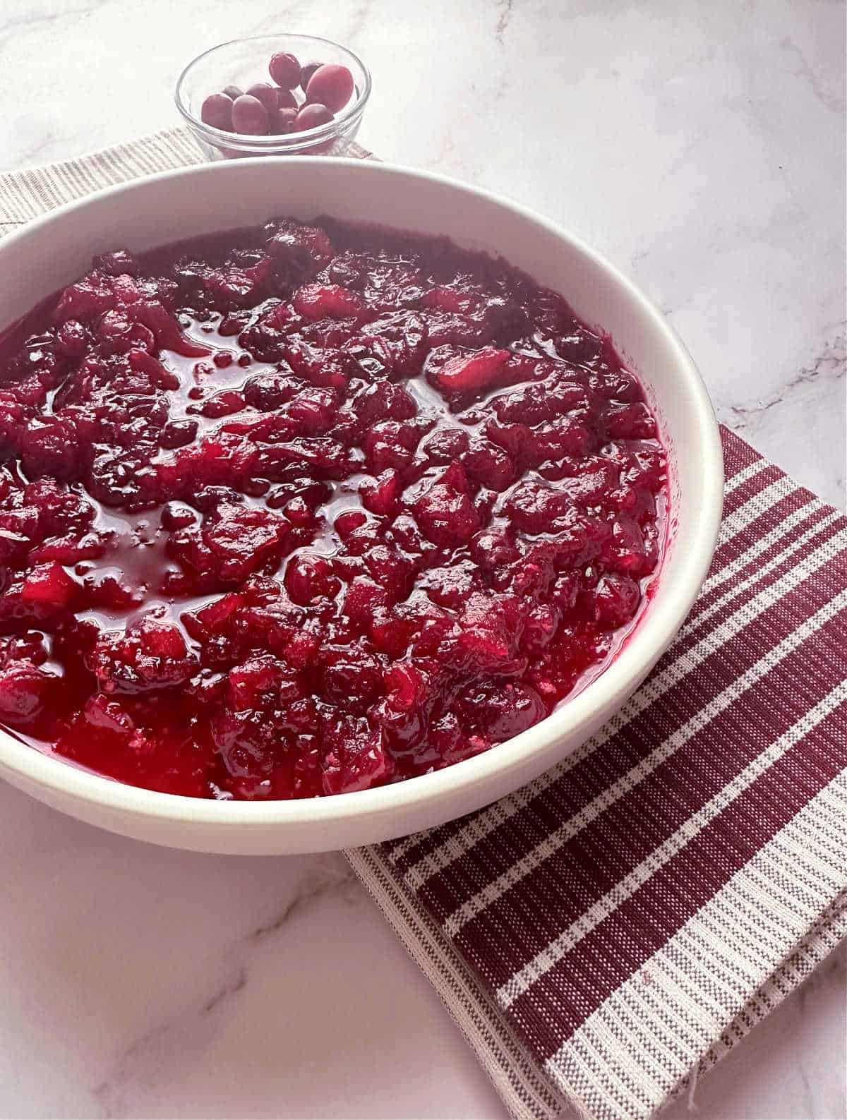 Cranberry sauce in a shallow white bowl with a burgandy and gray striped napkin underneath. 