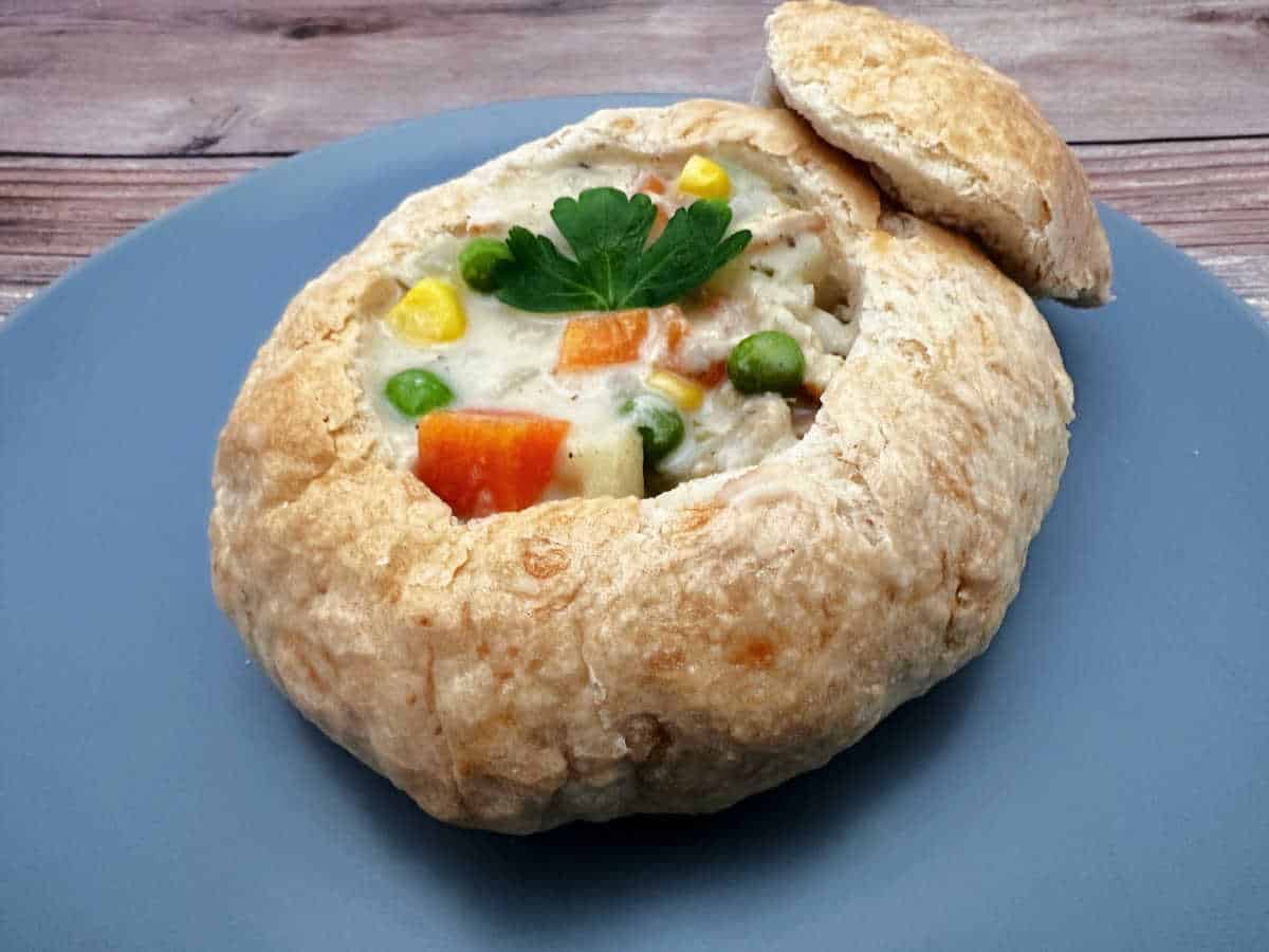 Soup, garnished with a sprig of fresh parsley, sits in a bread bowl on a dusty blue round plate.