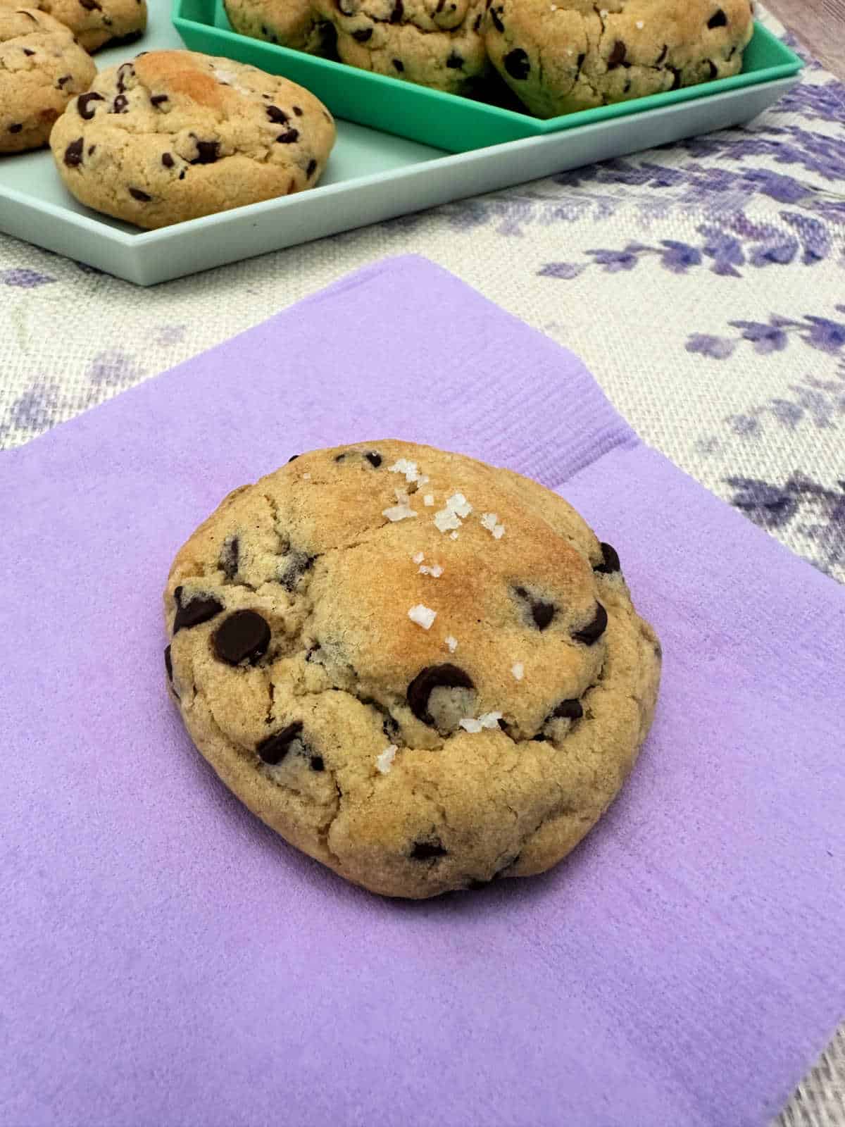 Cookie, sprinkled with salt, sits on a lavender napkin. Cookies in background are on a mint green platter resting on a lavender printed cloth.