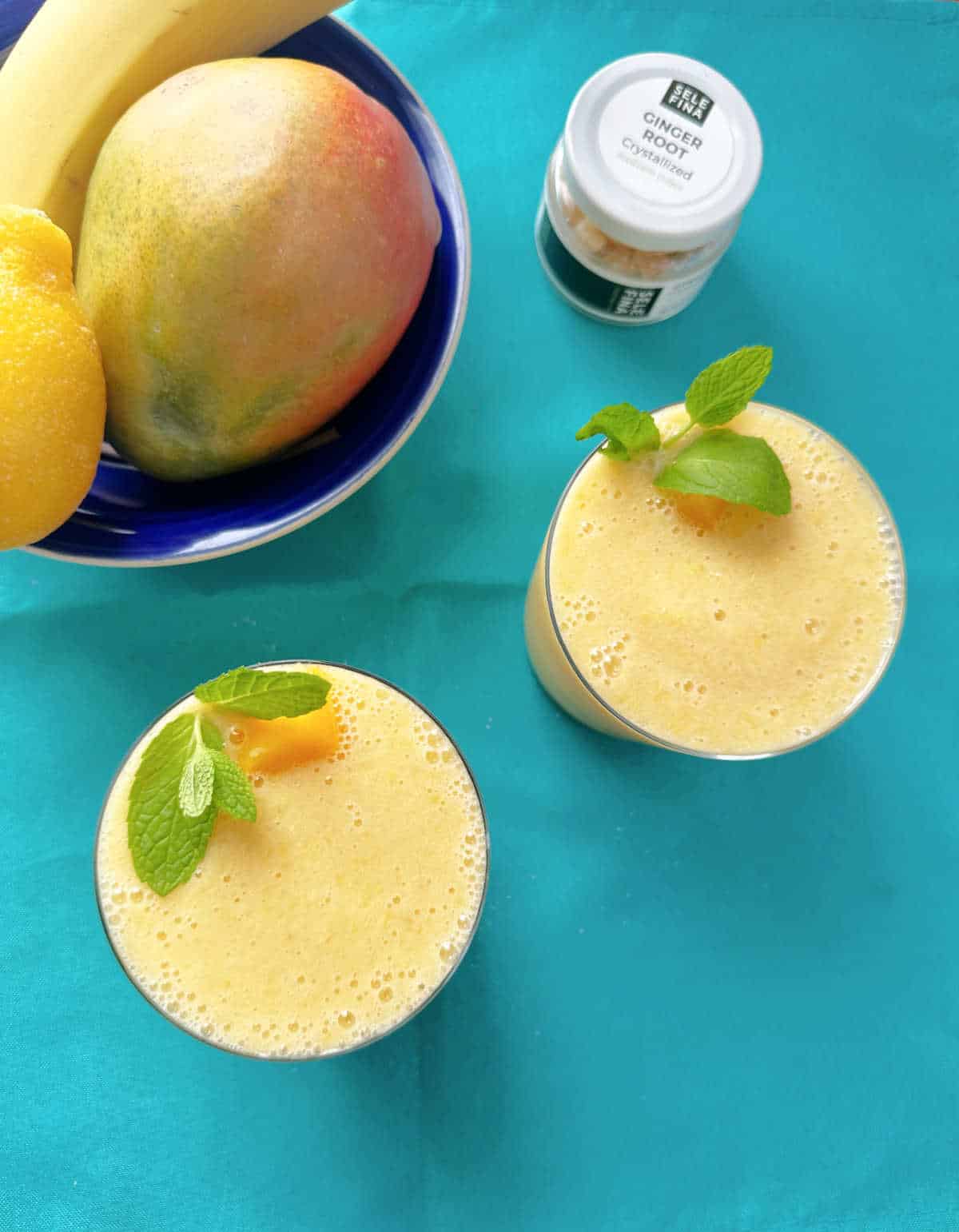 Overhead image of smoothies in short glasses on a teal cloth napkin. A bowl of fruit featuring the ingedients of the recipe sits to the top left. The yellow smoothies are garnished with mint leaves and mango pieces. 