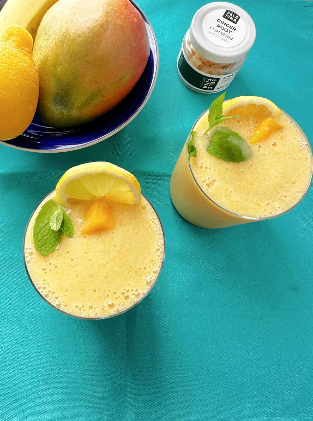 Overhead image of yellow smoothies in short glasses on a teal cloth napkin. Smoothies are garnished with lemon slices, mango pieces and fresh mint. Dark blue bowl of fruit - mango, lemon, banana. sits off to the side. 