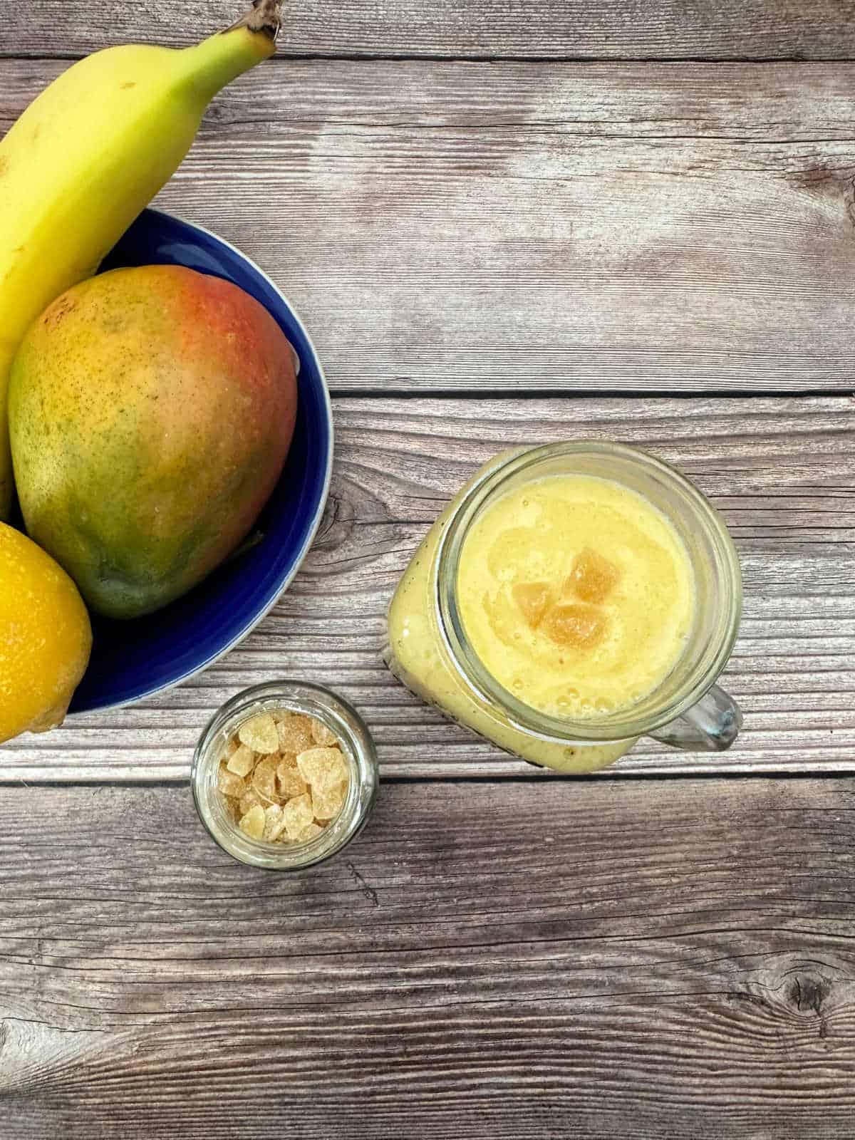 Dark blue fruit bowl holds a mango, lemon and banana. Smoothie sits in a mason jar on a wooden background. Open jar of crystalized ginger sits next to it. 