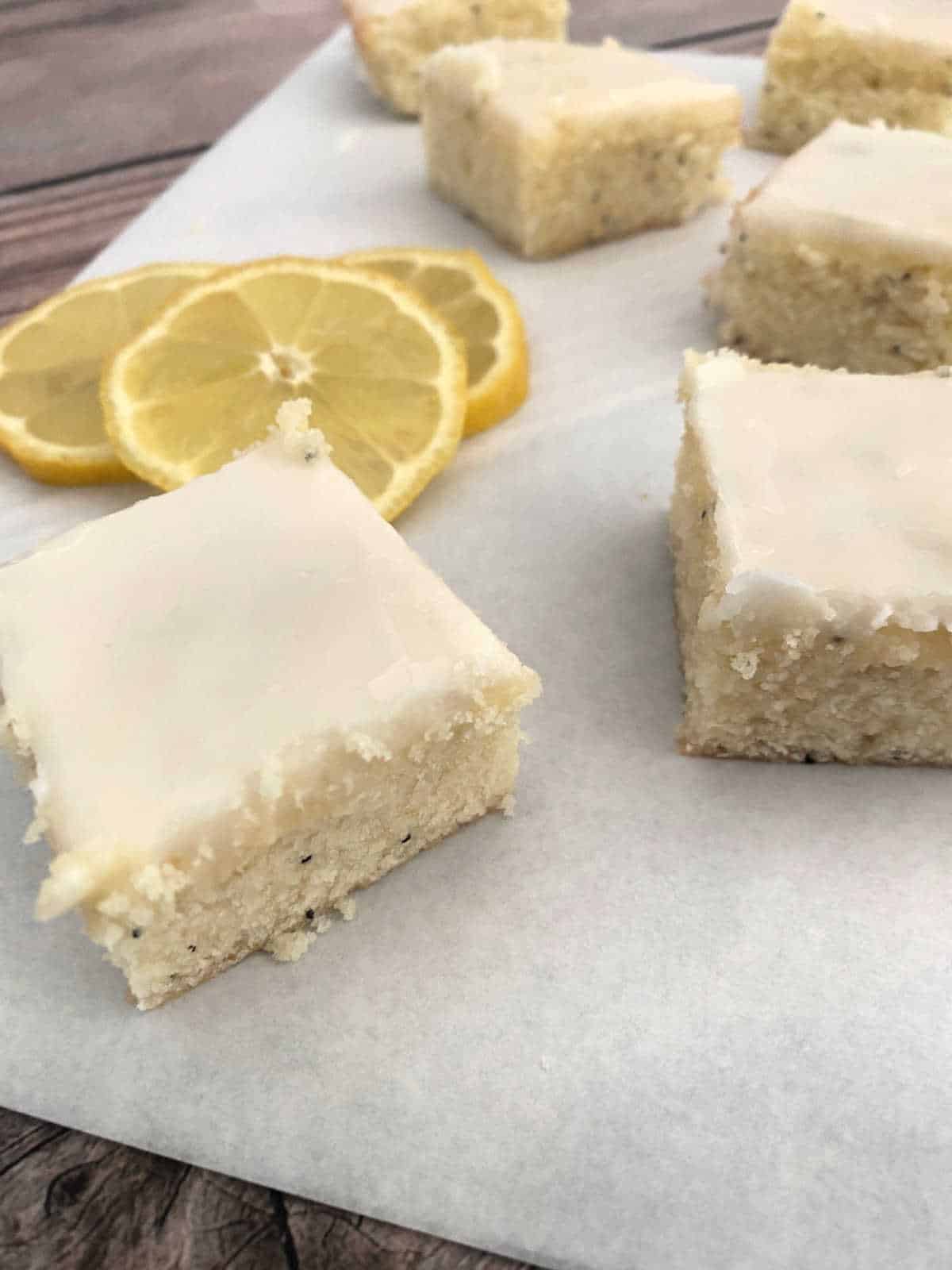 Bars sit on a sheet of parchment paper, surrounded by lemon slices, on a wooden background. 