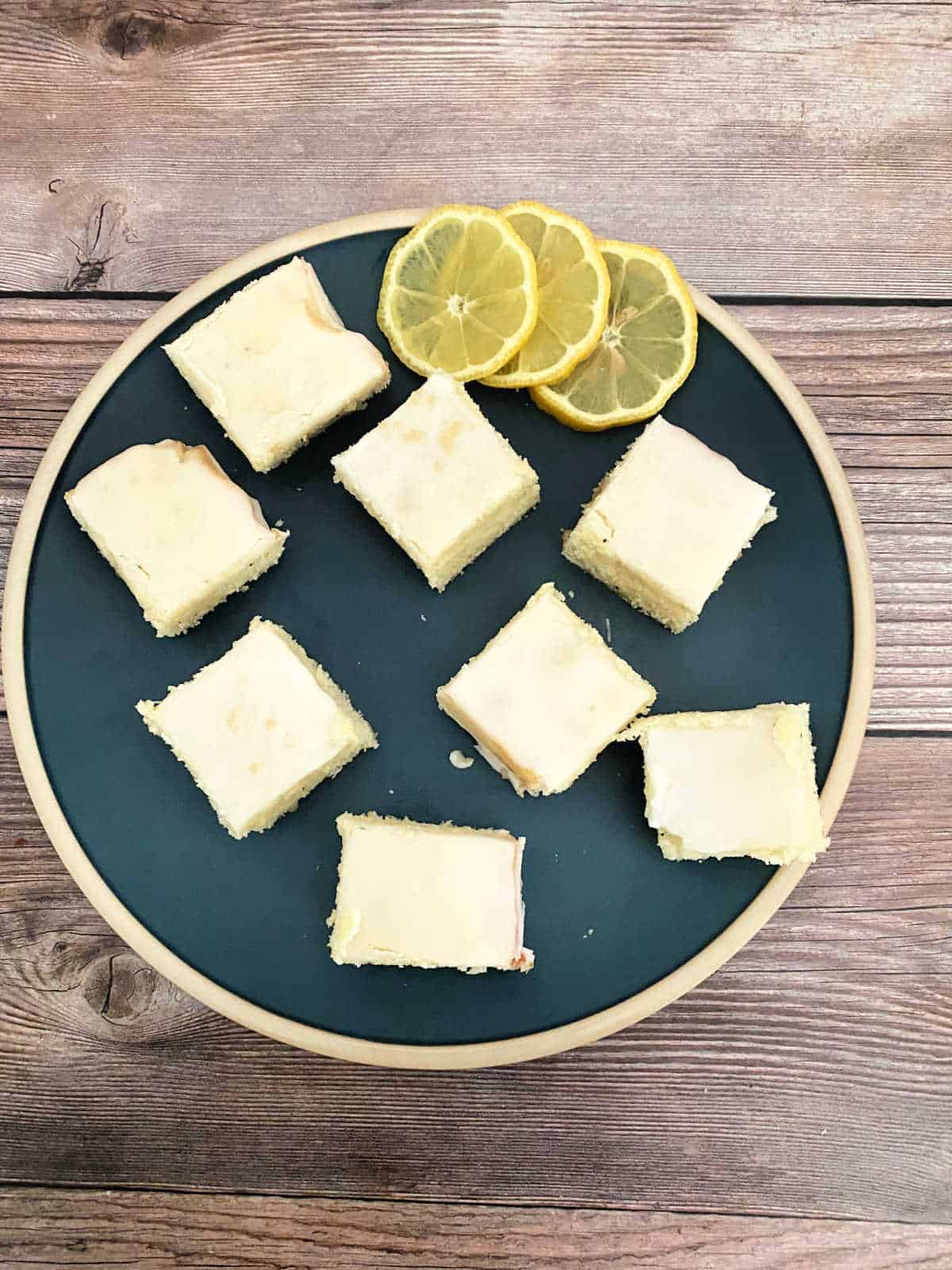 Lemon bars sit on a dark blue plate with lemon slics to the side. 