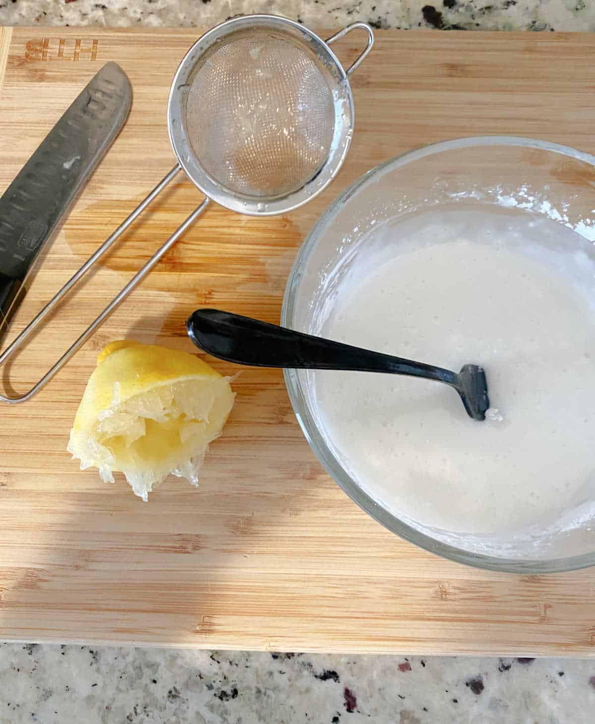 Process shot - on a wooden cutting board is a mesh sieve, a squeezed lemon, a knife and the powdered sugar and lemon glaze mixed together in a glass bowl. 