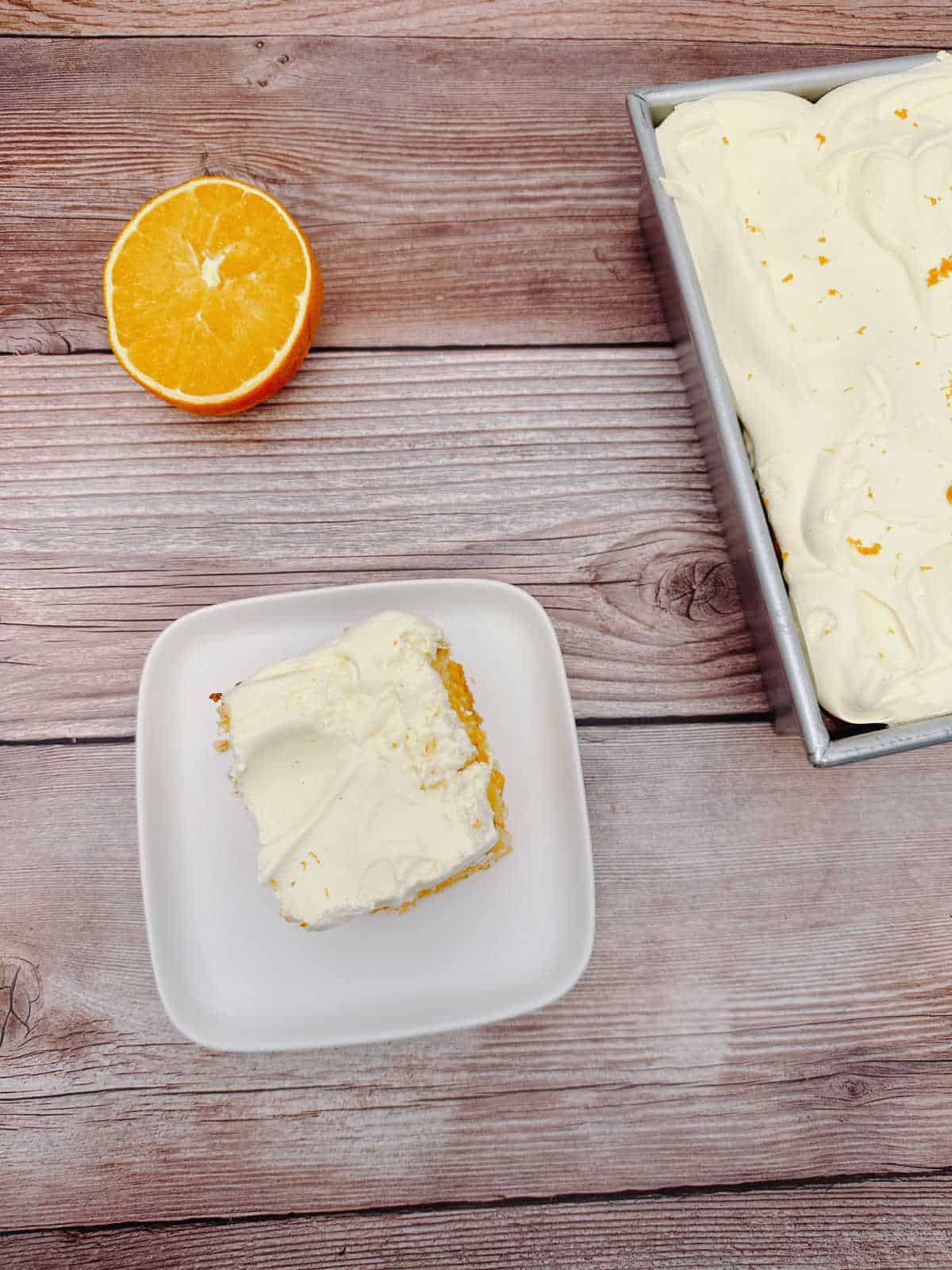 Overhead image of cake slice of a square plate. Cake in pan sits to the right, halved orange sits above the cake square. 