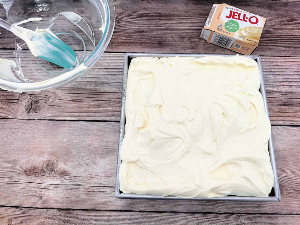 Process shot - Frosted cake sits on a wooden background next to the empty bowl of frosting. 