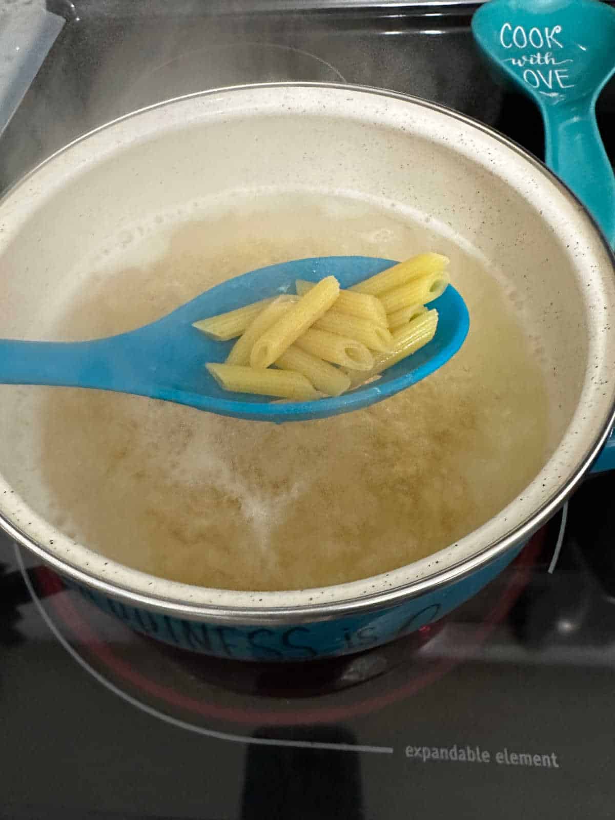 Process shot - slotted spoon holding up penne pasta over a pot of boiling water. 