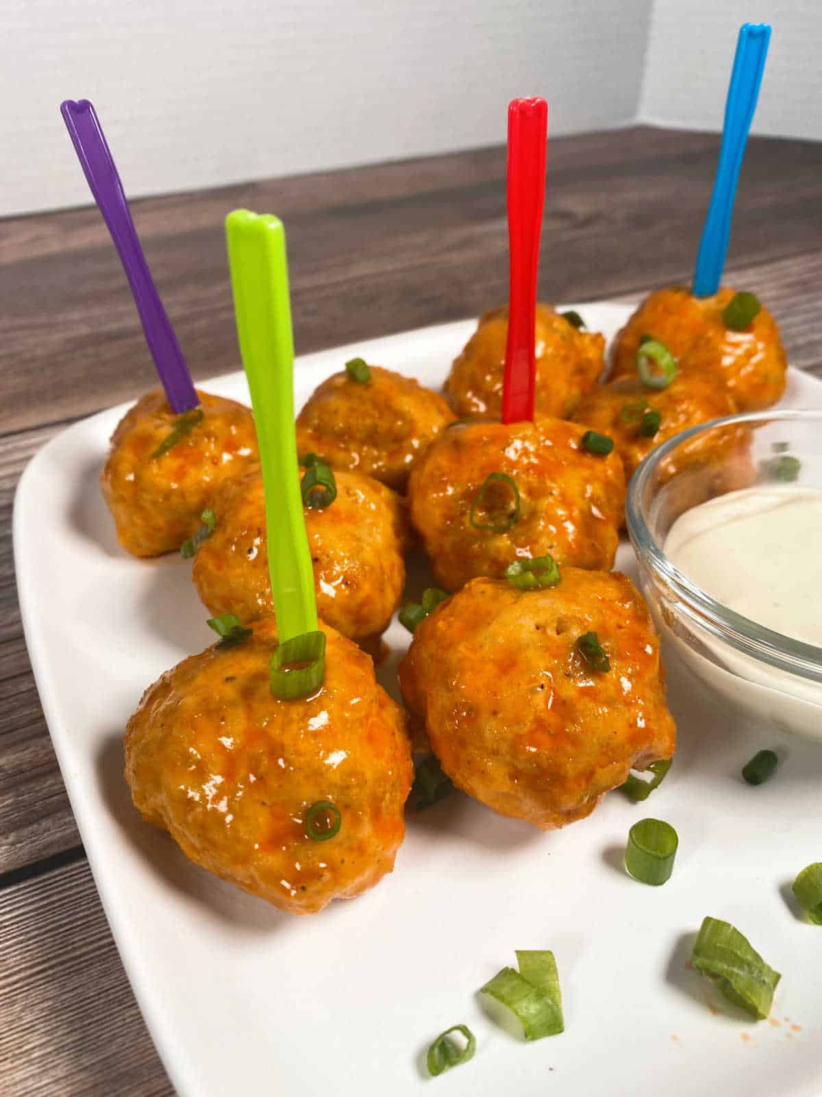 Close up image of meatballs on a white plate, garnished with chopped green onions. Meatalls have mini forks stuck in the tops of them. 