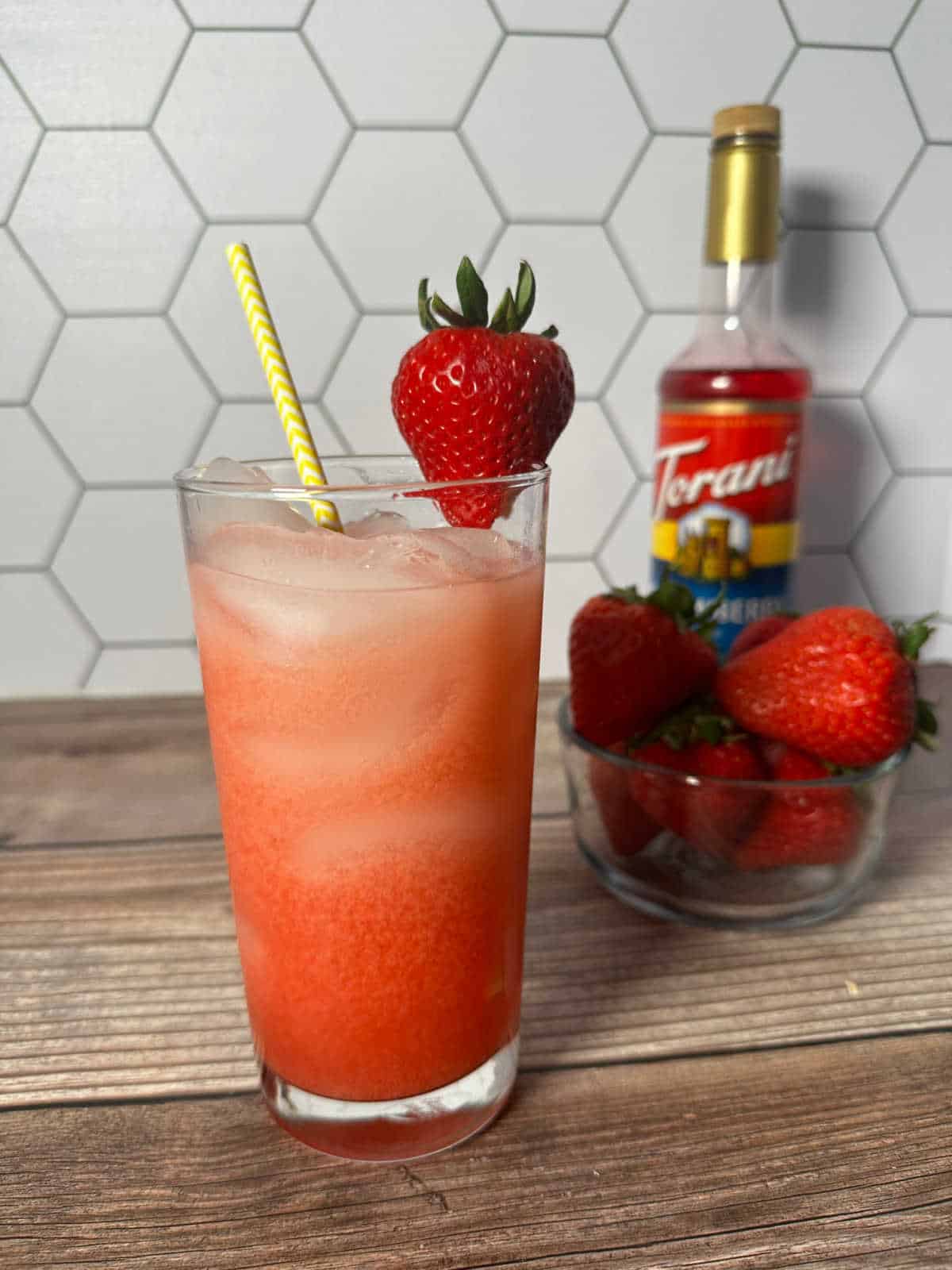 Tall glass with a yellow straw and strawberry garnish on top. Bowl of strawberries and the syrup sit in the background.