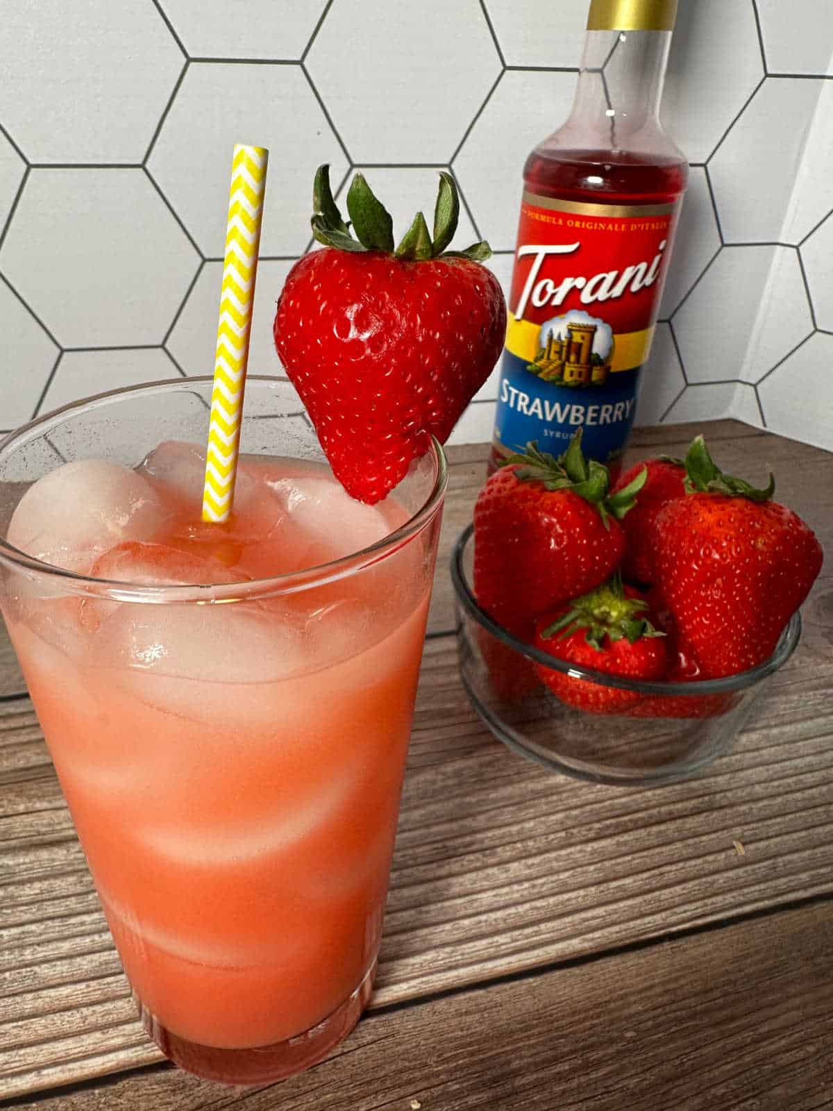 Close up of drink with yellow straw and strawberry garnish on a wooden backround with bowl of strawberries next to it. 