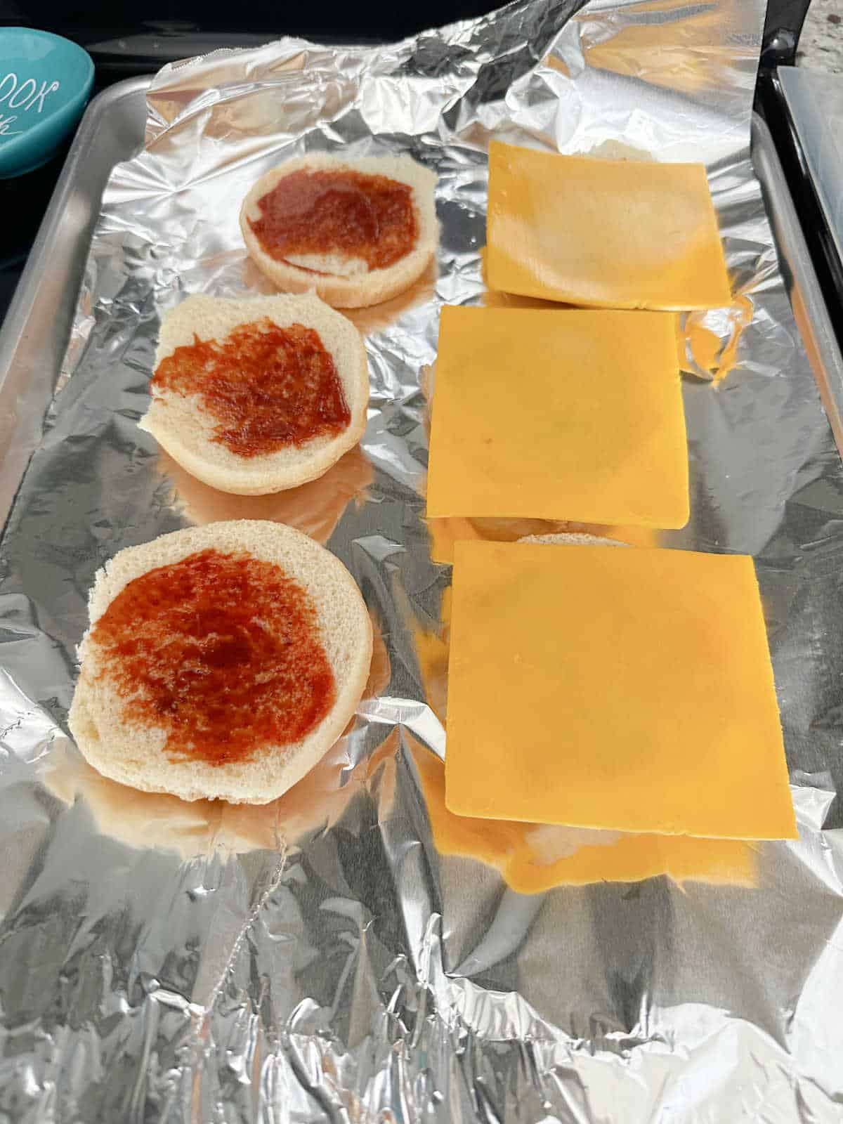 Burger buns with top half brushed with BBQ sauce and bottom half with a slice of cheese waiting to be toasted.