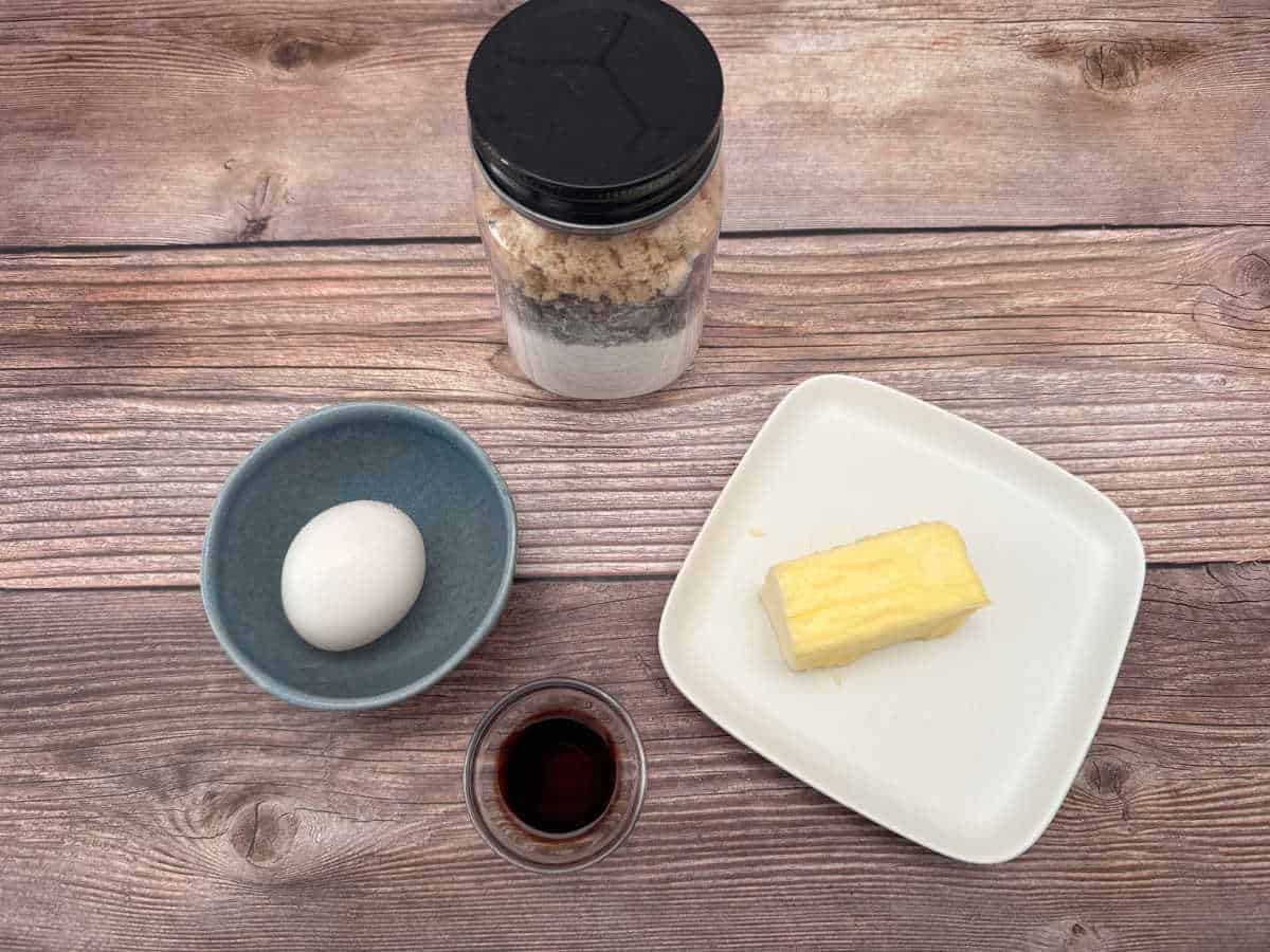 Cookie mix jar, egg in a blue bowl, unsalted butter on a white plate, vanilla extract in a tiny bowl on a wooden background. 